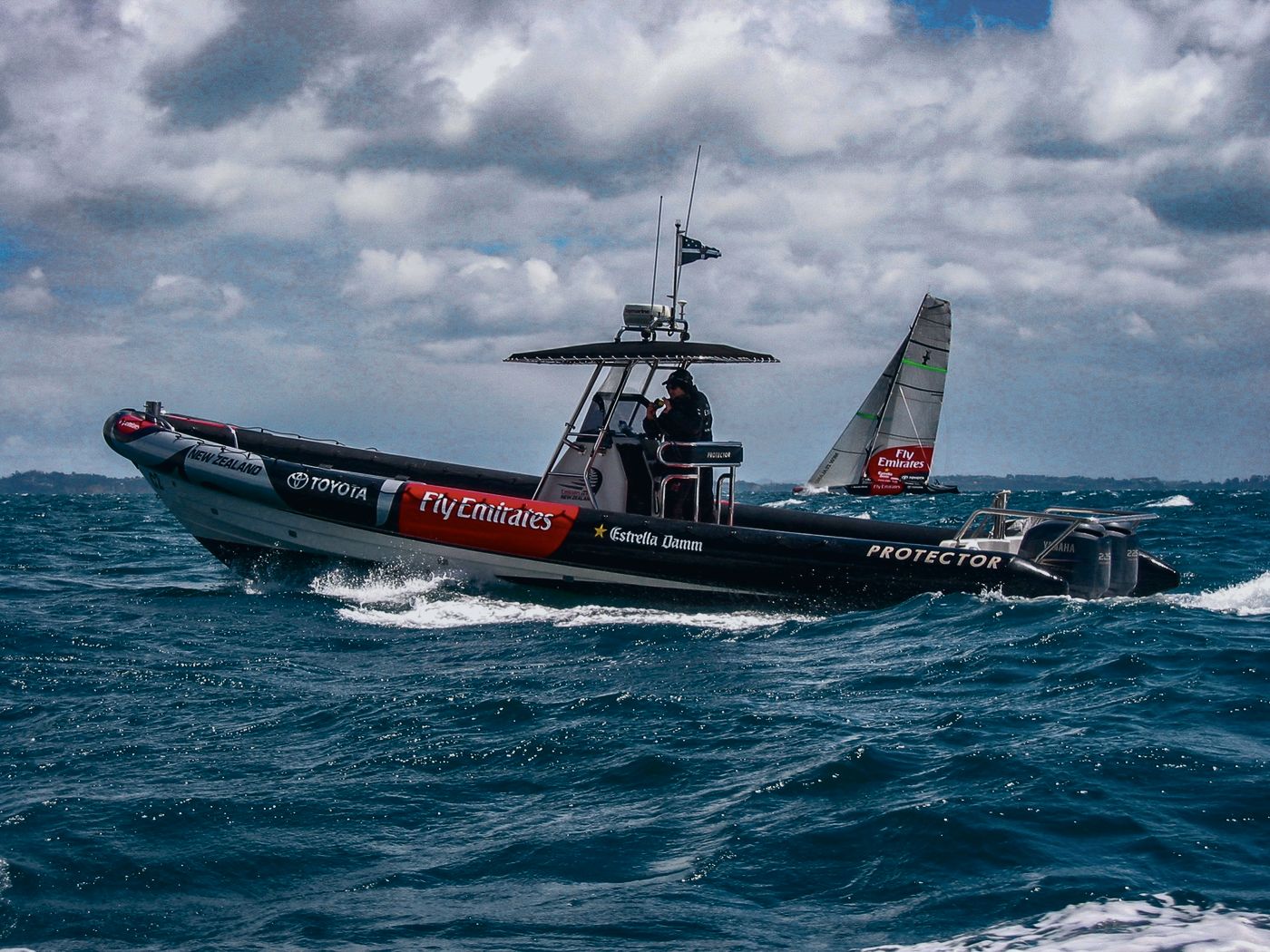 Protector chase boat in Emirates Team New Zealand livery at speed, with an America's Cup yacht behind